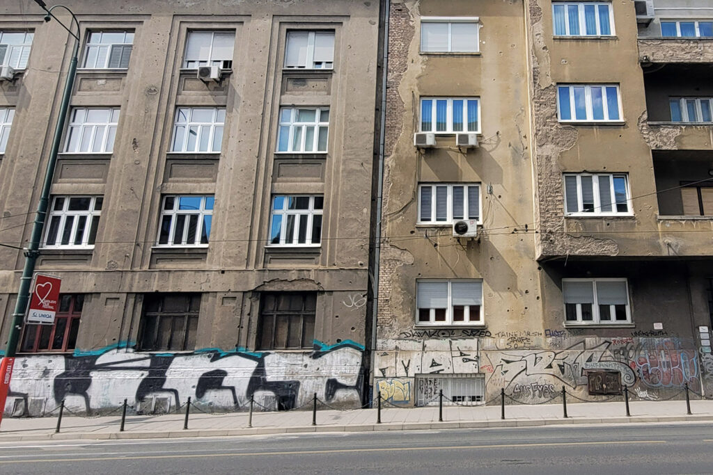 Spuren der Belagerung: Einschusslöcher an einer Häuserfassade in einer belebten Straße Sarajevos - City-Check Sarajevo, FemNews.de Hausfassade mit Einschusslöchern in Sarajevo